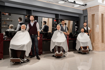 Barbers standing near clients covered with capes and sitting in hairdresser chairs.