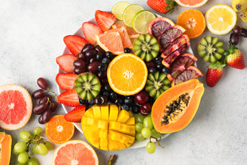 Raw fruits berries platter, mango, oranges, kiwi strawberries, blueberries grapefruit grapes on the white plate, on the off white table, top view, copy space