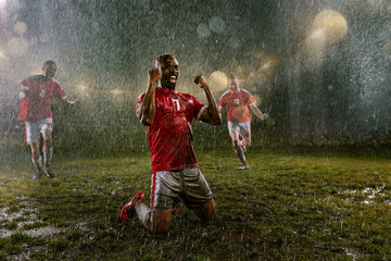 Soccer players on professional soccer night rain stadium. Three dirty players in rain drops emotionally rejoices victory. Black man kneel and scream