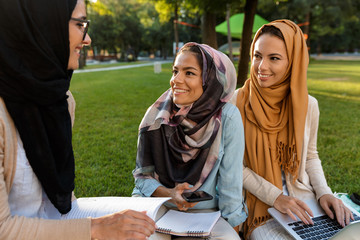 Happy young arabian women students using laptop computer and mobile phone outdoors in park.