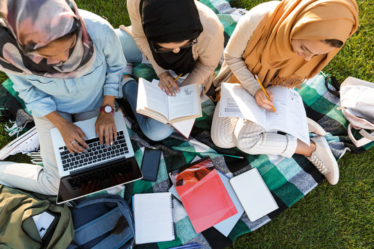 Women students using laptop computer and holding books in park.