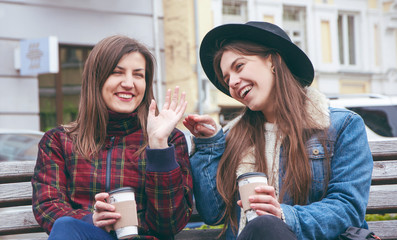A beautiful European girls drink coffee and chat on the bench against the backdrop of the city