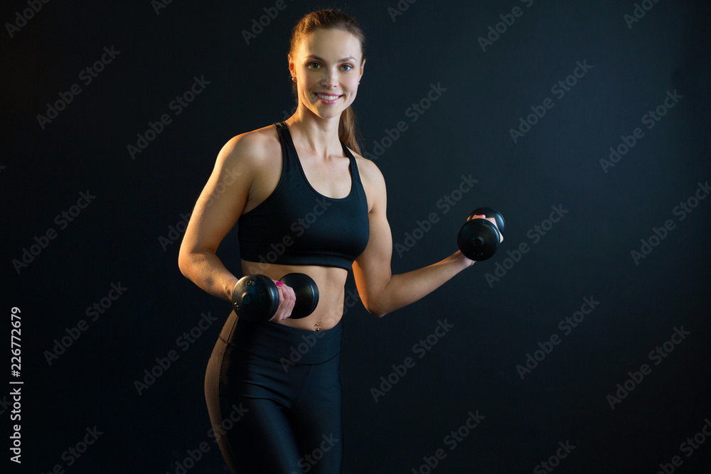 Wall mural beautiful young girl goes in for sports with dumbbells on a black background