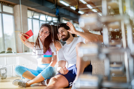 Picture of sporty beautiful fit couple sitting in bright gym and taking photo of them self. Smiling and looking at telephone.