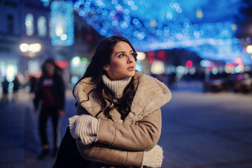 Cute young girl in winter coat standing in street and enjoying beautiful winter evening in decorated streets.
