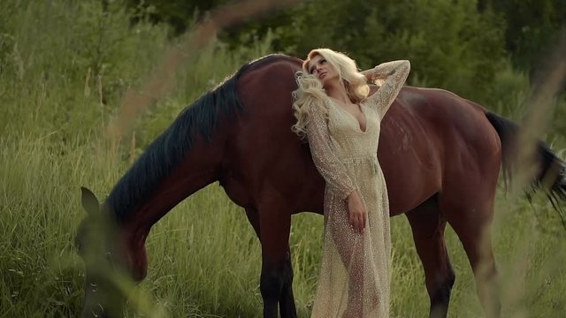 Enchanting slim graceful girl is posing with horse on meadow. Woman is leaning on horse flank animal is eating grass from lawn