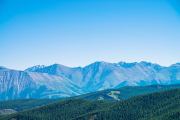 Giant mountains and glaciers above hills with forest. Snowy ridge under blue clear sky. Snow summit...