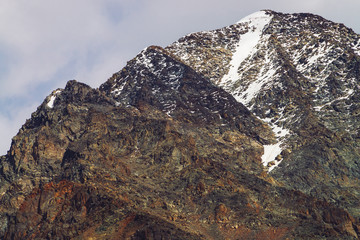 Snowy mountain top in cloudy sky. Rocky ridge under clouds. Overcast weather in highlands. Atmospheric minimalist landscape of majestic nature.