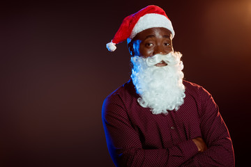 cheerful african american man in santa hat and beard posing with crossed arms on black with backlit