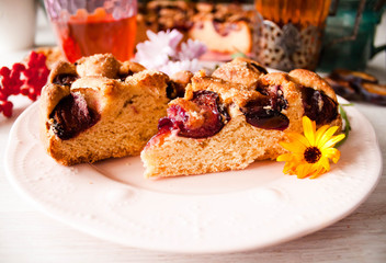 Homemade sweet round plum cake New York Times cuts on plate with cups, glasses of tea, coffee, flowers at back side on holyday Breakfast, birthday party on white table country style. Selective focus.