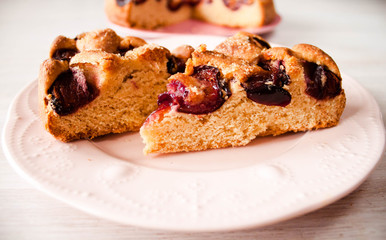 Homemade sweet round plum cake New York Times cutted on plate with cups, glasses of tea, coffee at back on holyday Breakfast or birthday party on white table country style. Side veiw. Selective focus.
