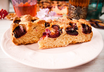 Homemade sweet round plum cake New York Times cutted on plate with cups, glasses of tea, coffee at back on holyday Breakfast or birthday party on white table country style. Side veiw. Selective focus.
