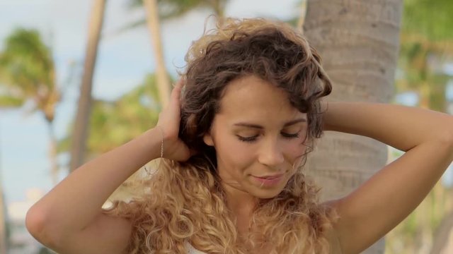 Cheerful young woman is standing alone in a tropical park in windy day. She is touching her long curly hair and correcting her summer dress