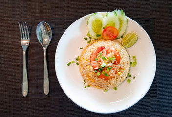Thai style shrimps fried rice garnished with sliced cucumber, tomato,lime and onion leaves served on the white plate with fork and spoon putted on the table cloth.