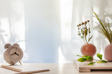 Photo of a desk with a concrete clock, an open notebook and vases with dried grasses on a background of sun-lit curtains