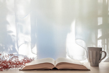 Photo of a desk with an open book, dried red grass and a gray cup with tea on a background of sun-lit curtains. Cpy space