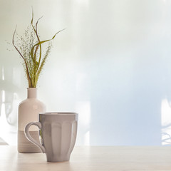 A photo of a desk with a white-gray vase with dried grass and a gray cup with coffee on a background of sun-lit curtains. Copy space