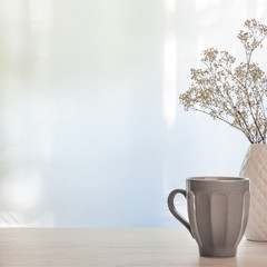 A photo of a wooden desk with a white vase with dried white flowers and a gray cup with tea on a background of sun-lit curtains. Copy space