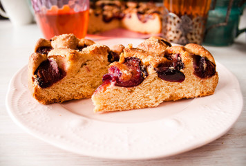 Homemade sweet round plum cake New York Times cutted on plate with cups, glasses of tea, coffee at back on holyday Breakfast or birthday party on white table country style. Side veiw. Selective focus.