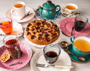 Homemade sweet round plum cake New York Times with cups, glasses of tea, coffee on saucers with spoons, forks, kettle on family Breakfast, birthday party on white country table. Selective focus