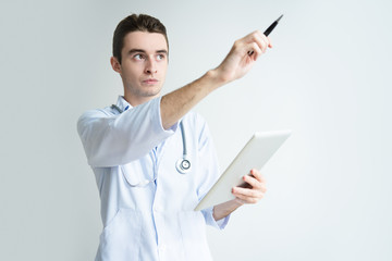 Serious male doctor holding tablet and pointing aside. Attractive guy standing and wearing white coat. Medicine and technology concept. Isolated front view on white background.