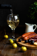 Aperitif concept. Wine pours into a glass goblet. On the table, meat appetizer, fried sausages,...
