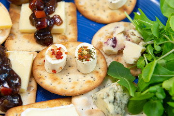 Crackers topped with a selection of cheeses, with sweet pickle and salad leaves. Close up view with focus on two oval shaped appetizer cheeses. On a blue plate.