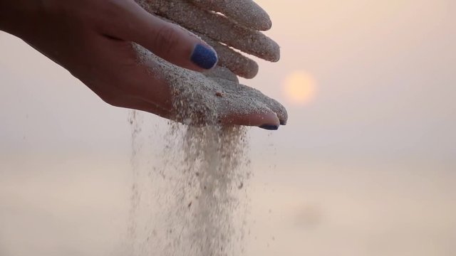 Arms of young woman with sand strewed or falling from it. Female hand pouring sea sand through her fingers at sunset against an ocean background. Grit drizzling from fist of girl. Slow motion Close up