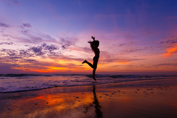 Silhouette of happy joyful woman jumping and having fun on the beach against sunset. The concept of freedom and relaxation. Reflection of the amazing sunset sky. The sky is in incredible clouds.