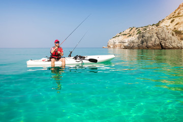 Man fishing on a kayak in the sea with clear turquoise water. Fisherman kayaking in the islands. Leisure activities on the ocean. - obrazy, fototapety, plakaty