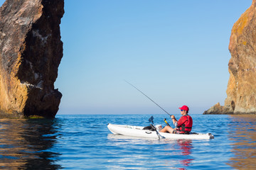 A man fishing on a kayak boat in the sea near the rocks at the shore of island mountain. Fisherman...