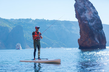 A man fishes on a fishing tackle in the standup paddleboard. Young fisherman on the SUP surfing in the sea near the island with rocks and mountains.