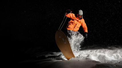 Active sportsman snowboarder in orange sportswear and mask jumping on a snowy hill at night