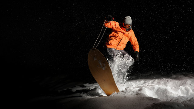 Active sportsman snowboarder in orange sportswear and mask jumping on a snowy hill at night
