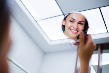 My teeth look perfect! Beautiful young woman looking at mirror with smile in dentist’s office