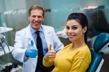 Confidence in your dentist. Cheerful doctor and his beautiful patient looking at camera with smile and gesturing thumb up in dentist’s office