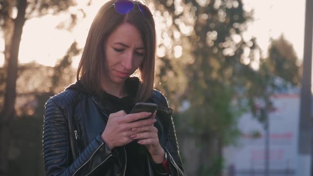 Attractive young girl stands in middle of busy buzzling city on summer sunset. Gorgeous young woman in a bright sunlight uses her phone.