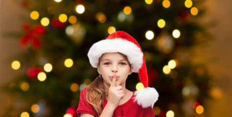 Fototapeta na wymiar holidays and children concept - happy girl in santa hat making shh gesture over christmas tree lights background