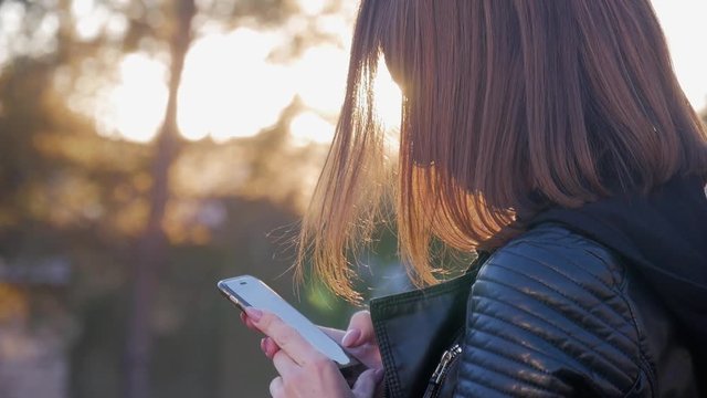Attractive young girl stands in middle of busy buzzling city on summer sunset. Gorgeous young woman in a bright sunlight uses her phone.