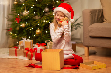 Obraz na płótnie Canvas christmas, holidays and childhood concept - smiling girl in santa helper hat with gift box and teddy bear at home