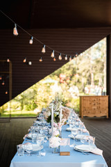 Long festive table with blue tablecloth decorated with flowers and candles under garland