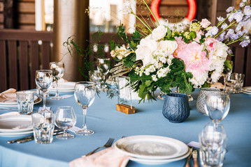 Festive round table with a blue tablecloth, dishes, wine glasses and a bouquet of flowers in the center