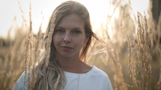 Attractive young woman with long blond hair wearing white t shirt is standing in meadow on sunny day and looking at camera. Handheld slow motion medium shot