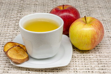 Close-up view of a cup of apple tea with two apples and dehydrated slices of the fruit with a patterned table cloth in the background