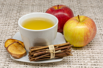 A cup of apple tea with two apples and dehydrated slices of the fruit with a patterned table cloth in the background and a bunch of cute little sticks