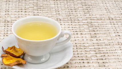 A cut view of a cup of apple tea with two dehydrated slices of the fruit with a patterned table cloth in the background