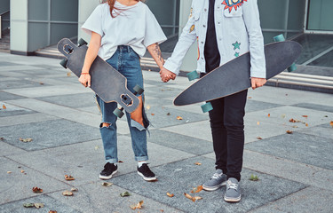 Cropped photo of young trendy dressed skaters hold hands while standing near skyscraper.