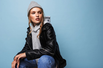 Young beautiful woman with wavy hair in gray hat and black leather jacket thoughtfully looking aside over blue background isolated