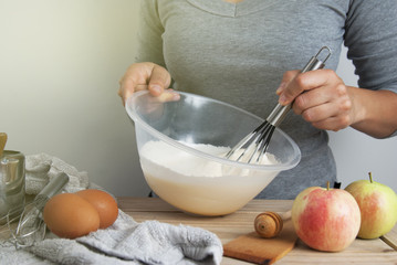 Close-up young woman's hands holding whisk and bowl while making pie, cake. Female cooking dough for pie on wooden table. Preparring dessert.