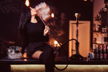 Sexy brunette girl in seductive black clothes smokes a hookah while sitting on counter in a nightclub or bar.